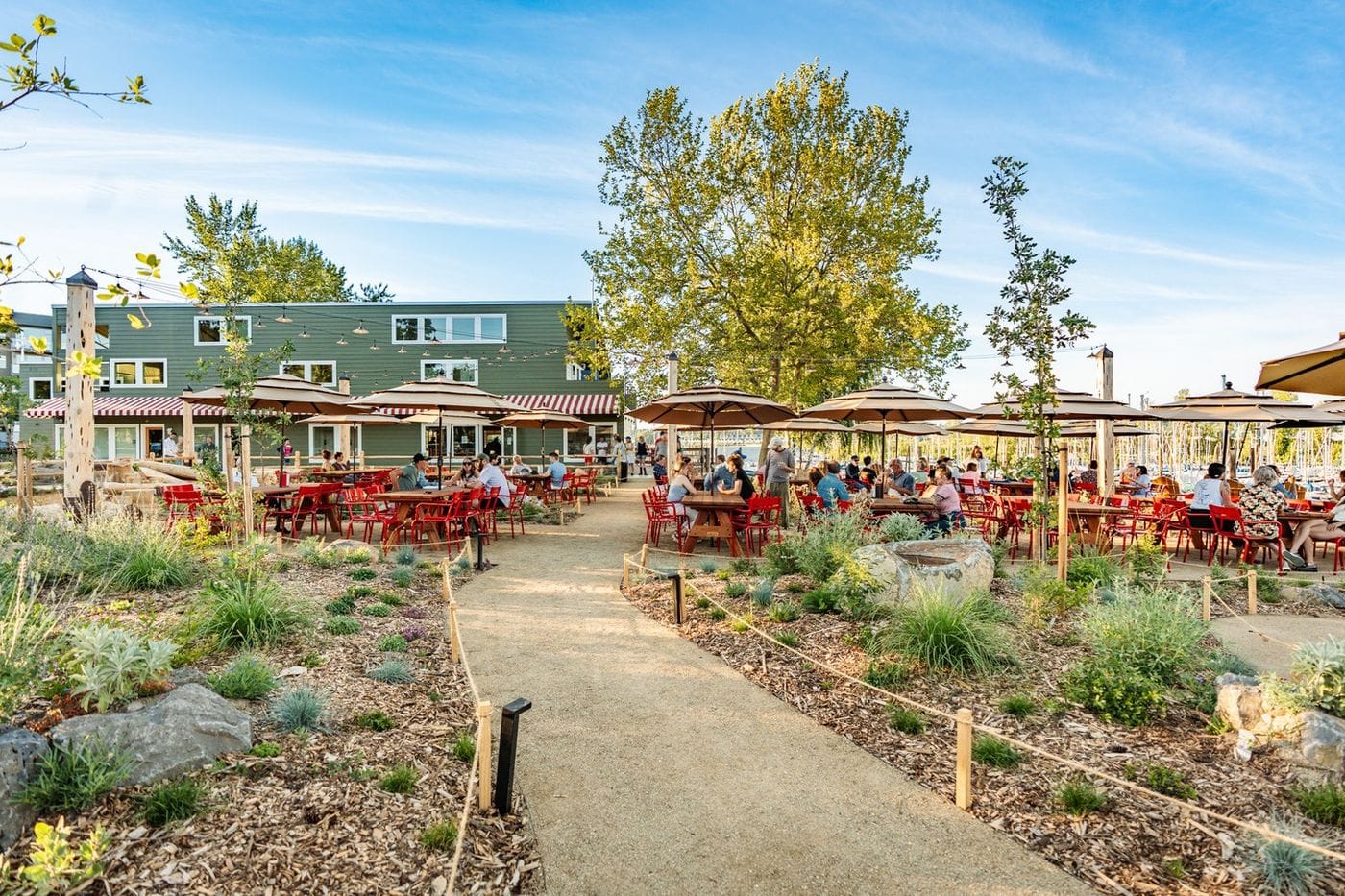 Outdoor waterfront dining area with red tables and umbrellas, guests seated along a landscaped path, and a marina with boats visible in the background on a sunny day.